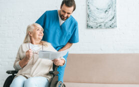 Smiling Young Male Nurse Looking At Happy Senior Woman Reading Newspaper In Wheelchair