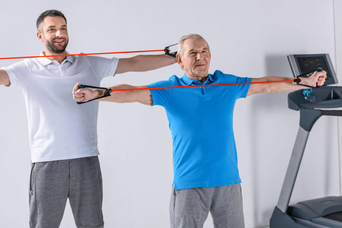 Smiling Rehabilitation Therapist And Senior Man Exercising With Rubber Tapes