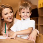 Happy Educator Hugging Adorable Boy At Table In Kindergarten