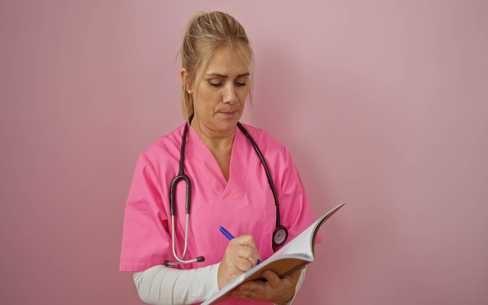 Woman,Nurse,Writing,Notes,In,A,Notebook,Wearing,Pink,Scrubs